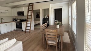 Un vaste espace de vie comprenant un coin repas avec table et chaises, une cuisine dotée de meubles blancs et d’appareils électroménagers, et une chambre en mezzanine accessible par une échelle.