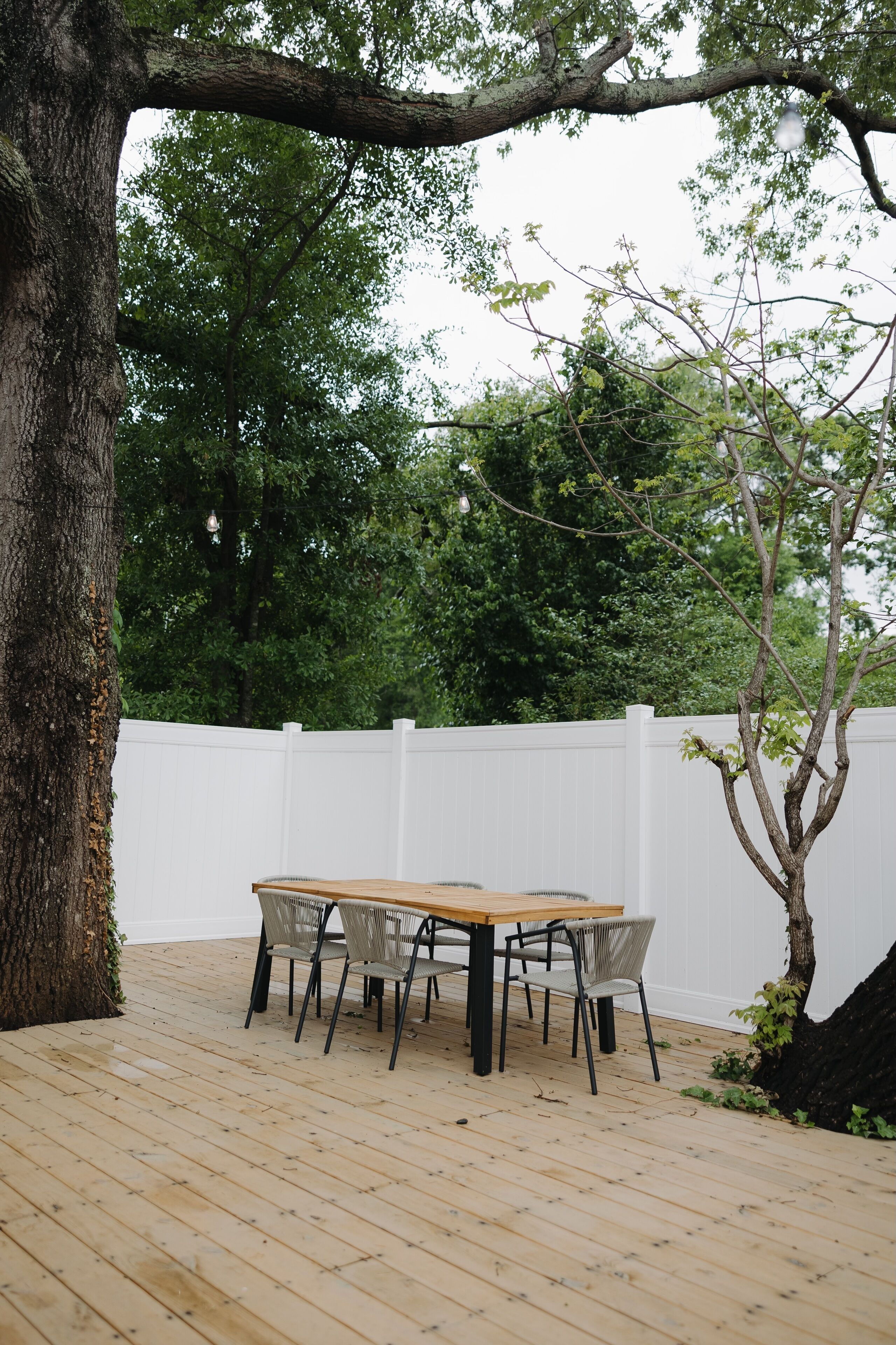 Una terraza de madera al aire libre con una mesa y sillas, rodeada por una valla blanca y árboles.
