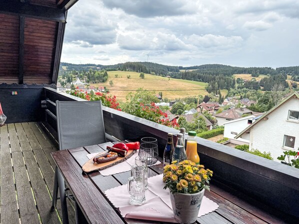 A balcony with a table set for breakfast, a chair, a potted plant, and a scenic view of a village and hills.