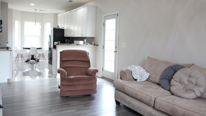 Amplia sala de estar con ventilador de techo, un sofá, un sillón reclinable y una cocina al fondo.