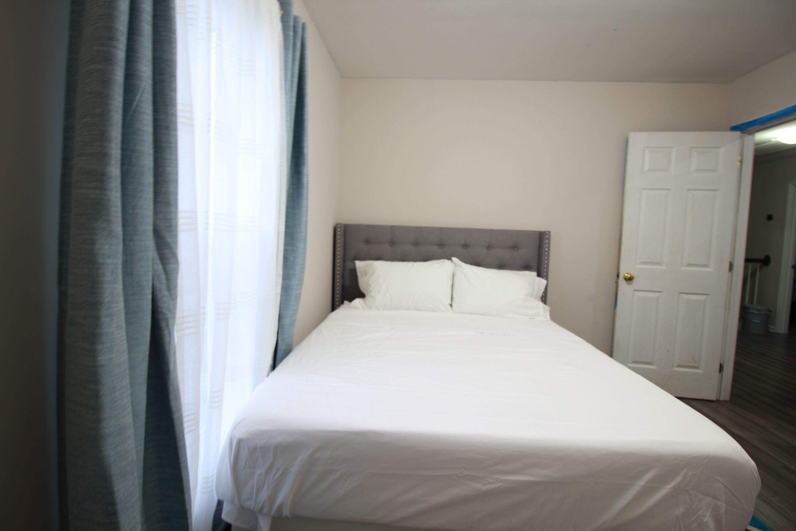 Een netjes opgemaakt bed met een grijze hoofdbord in een kamer met witte muren en een gesloten deur.