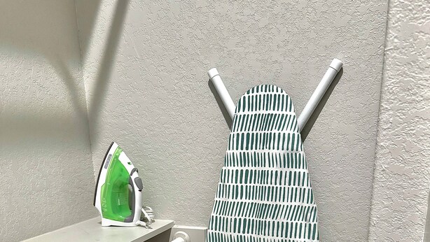 Rincón de una habitación con una tabla de planchar blanca, un planchador verde y blanco, y un estante blanco.