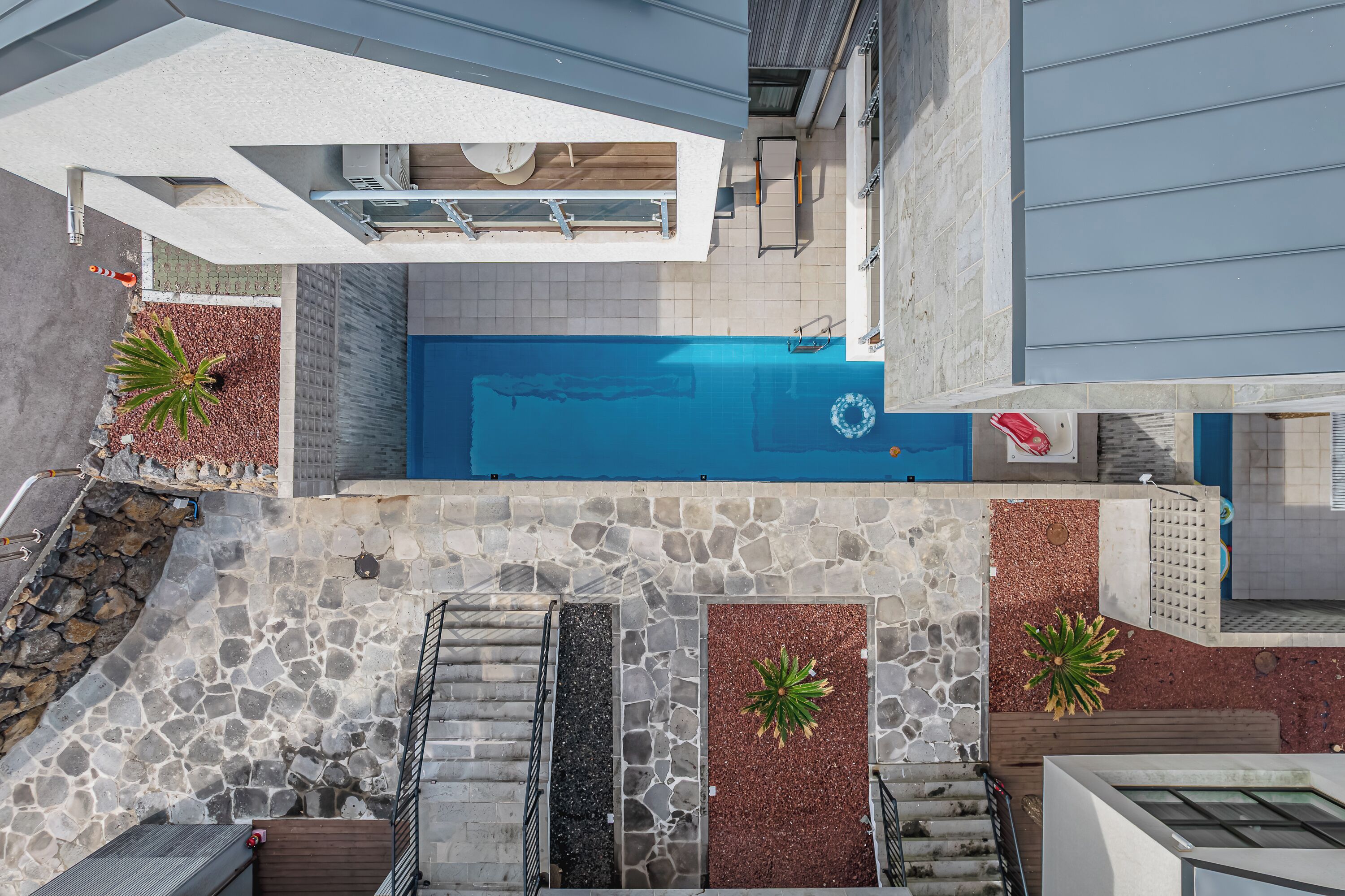A rooftop pool with stone and tiled flooring, surrounded by potted plants and a staircase leading down.