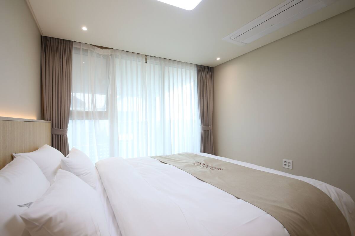 Ein Hotelzimmer mit zwei Betten, einem großen Fenster mit durchsichtigen Vorhängen, einer Decken-Klimaanlage und einem Lichtschalter an der Wand.