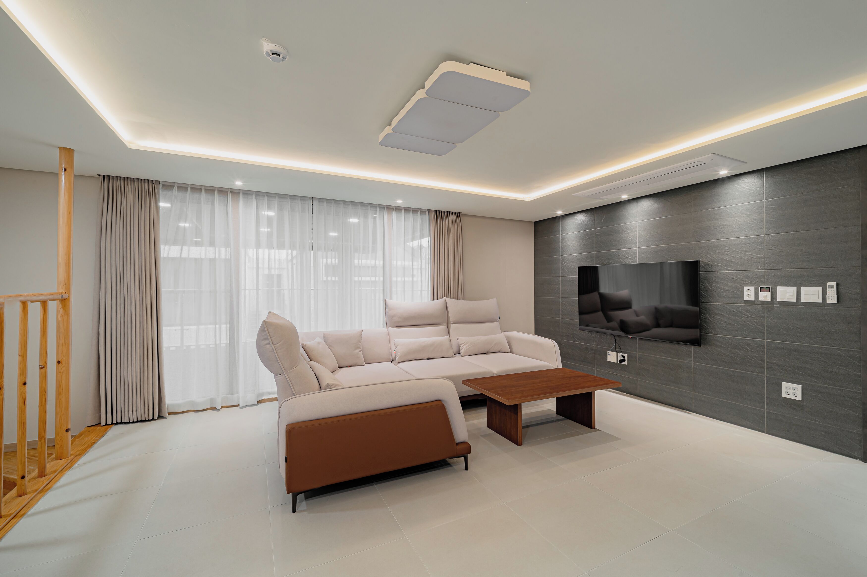 A modern living room with a beige sectional sofa, a wooden coffee table, a flat-screen TV mounted on a dark wall, and a staircase with wooden railings.