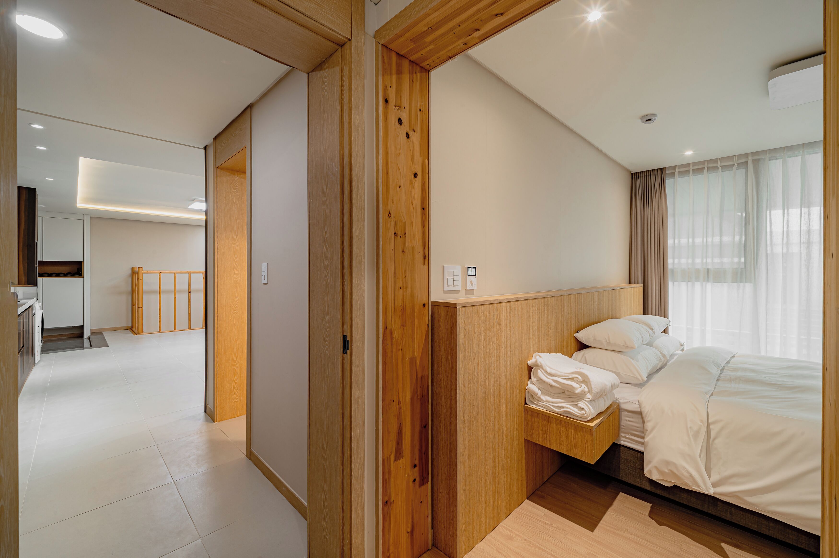 A modern hotel room with a wooden headboard, a neatly made bed, and a view of the city through large windows.