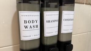 Drie doseerders voor douchegel, shampoo en conditioner bevestigd aan een betegelde muur.