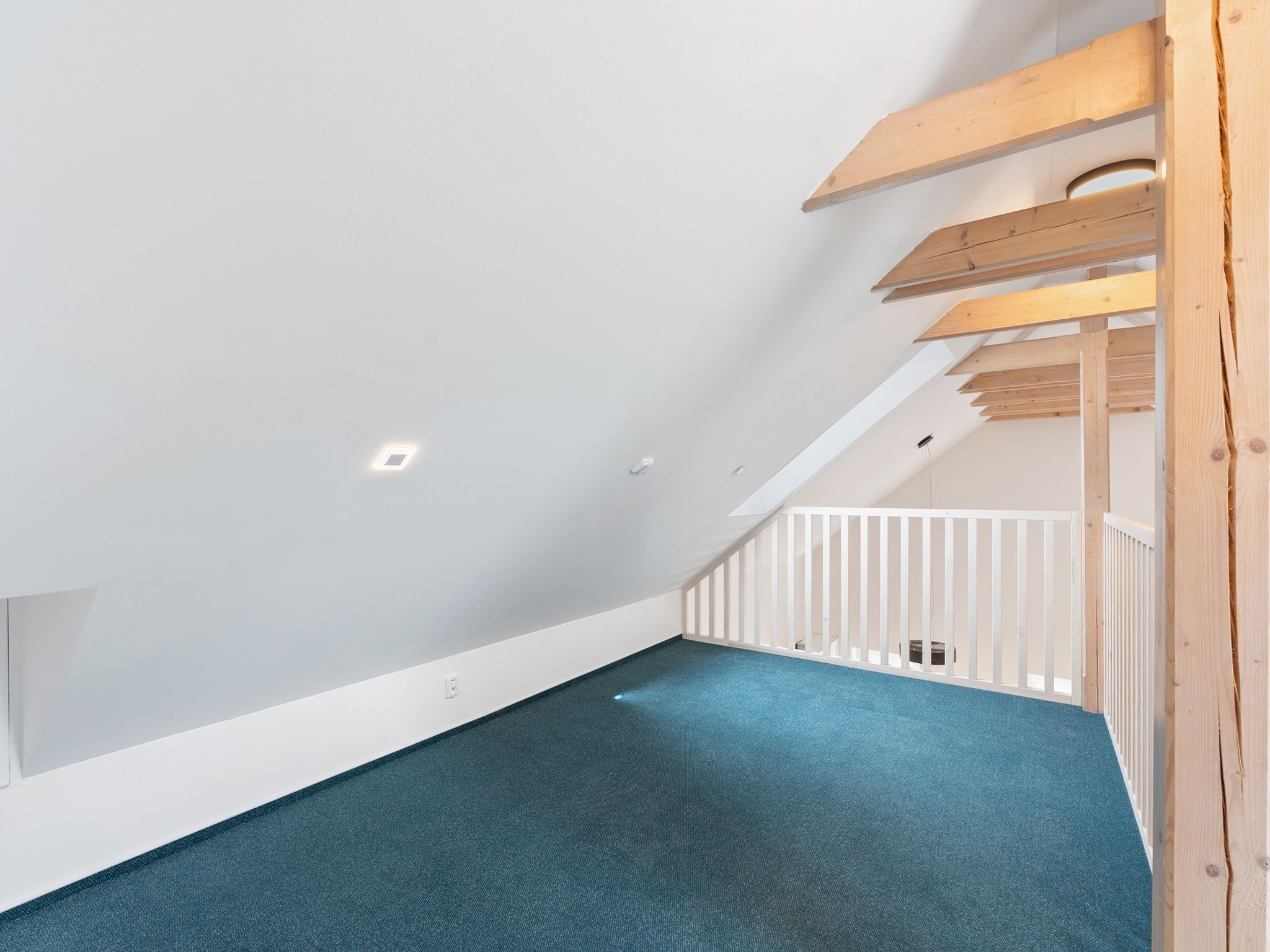 A spacious attic room with a sloped ceiling, wooden beams, and a blue carpet.