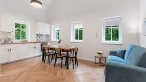 A modern kitchen with white cabinets, a wooden dining table, and a blue sofa in a bright room with large windows. - V kapradí by Interhome (Hřensko)