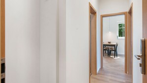 Un couloir avec un plancher en bois et des murs blancs. Une porte se trouve sur le côté droit et donne sur une pièce où se trouvent une table et des chaises.