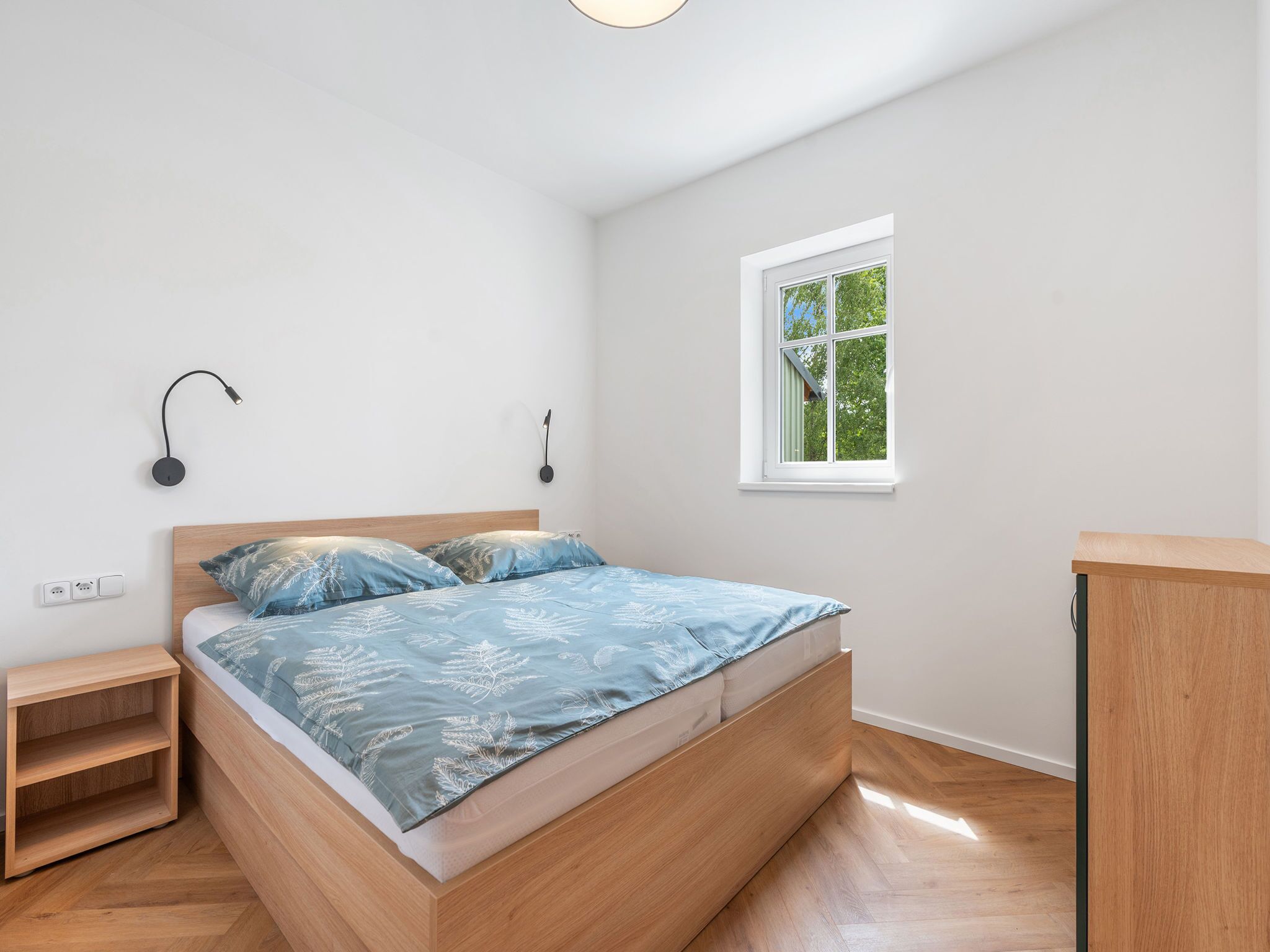 A bedroom with a wooden bed, a nightstand, a wall-mounted lamp, a window with a view of greenery, and a wooden desk.