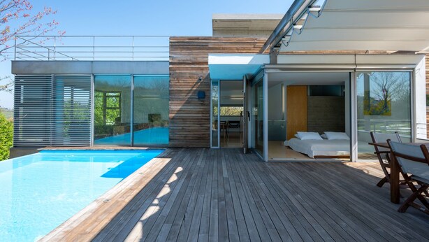 Et moderne hus med swimmingpool, træterrasse og glasdøre, der fører til et soveværelse.