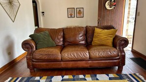 Un sofá de cuero marrón con tres cojines en una habitación con suelo de madera y una alfombra estampada.