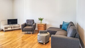 Ein modernes Wohnzimmer mit grauem Sofa, einem grauen Sessel, einem kleinen Hocker, einem Holztisch mit Blumenvase und einem Fernseher auf einem weißen Schrank.