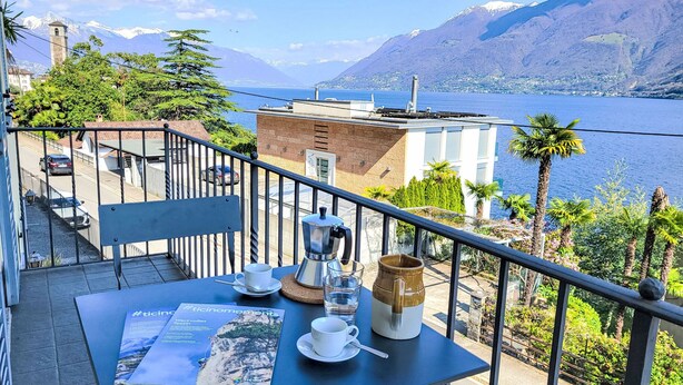 Ein Balkon mit Tisch für zwei Personen, einer Kaffeemaschine und einer Zeitschrift. Blick auf einen Berg und ein Gewässer.