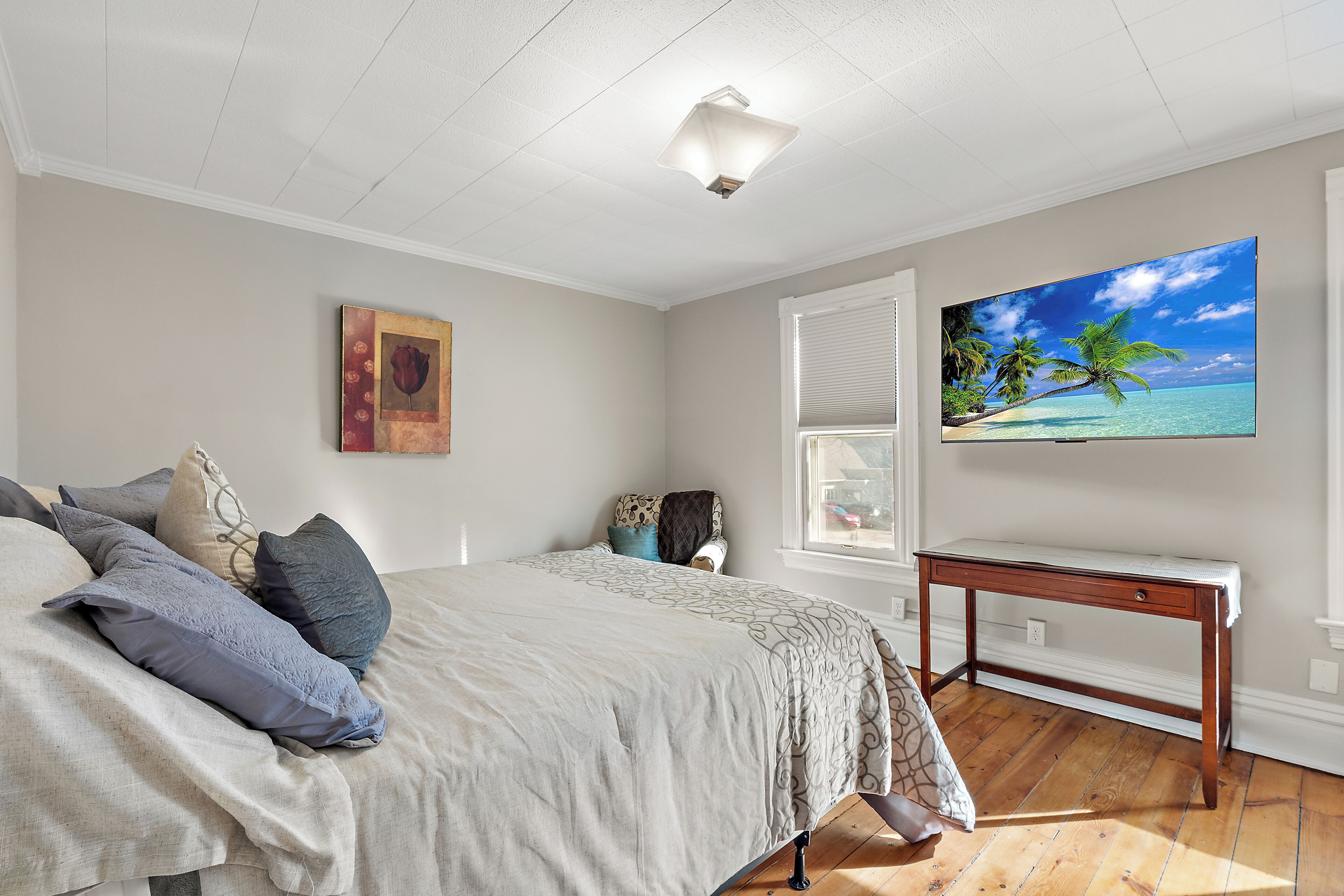 Une chambre à coucher avec un lit, un bureau, un tableau représentant une plage et une fenêtre avec des stores.