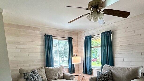 Uma sala de estar com dois sofás, uma poltrona marrom, um tapete estampado e um ventilador de teto.