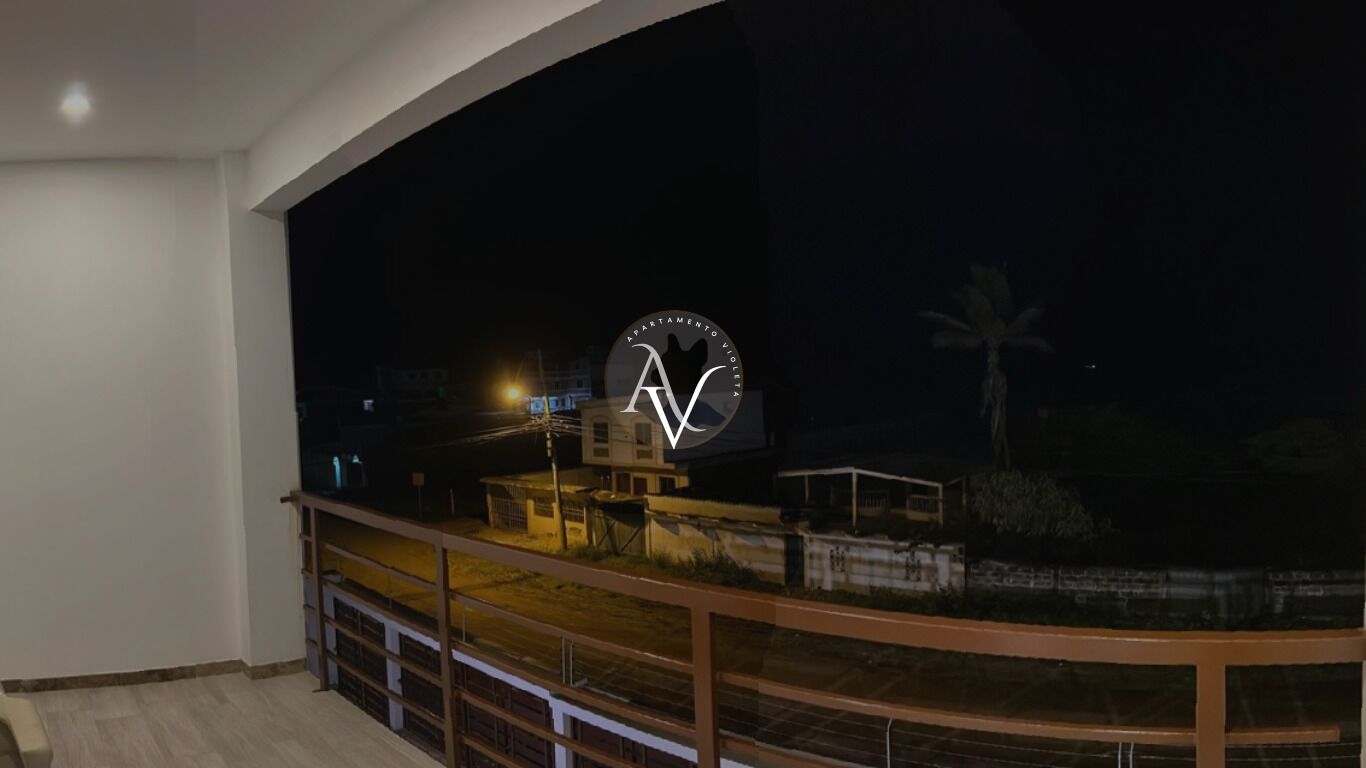 Vista nocturna desde un balcón con barandilla, con vista a una calle con edificios y una palmera.