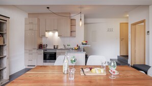 Une cuisine moderne avec des meubles en bois, une table à manger dressée avec de la nourriture et des boissons, et un petit îlot central sur lequel se trouve un panier de fruits.