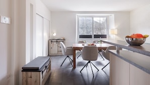 Une cuisine moderne avec une table en bois, des chaises, un plan de travail sur lequel se trouve un saladier rempli de fruits, et une fenêtre donnant sur des arbres.