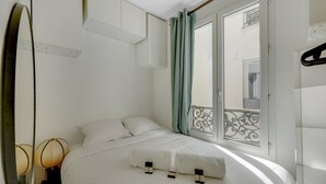 A neatly made bed in a room with a large window, a mirror, and a wall-mounted shelf.