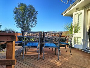 A wooden deck with two metal chairs and a bench, a white building with a window, and a tree in the background. -  Country Cottage Delight in the Hurunui.
(Spotswood/Cheviot)
 (Spotswood)