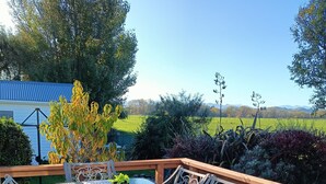 A deck with a glass-top table and blue cushioned chairs, overlooking a green landscape with a blue building in the background. -  Country Cottage Delight in the Hurunui.
(Spotswood/Cheviot)
 (Spotswood)