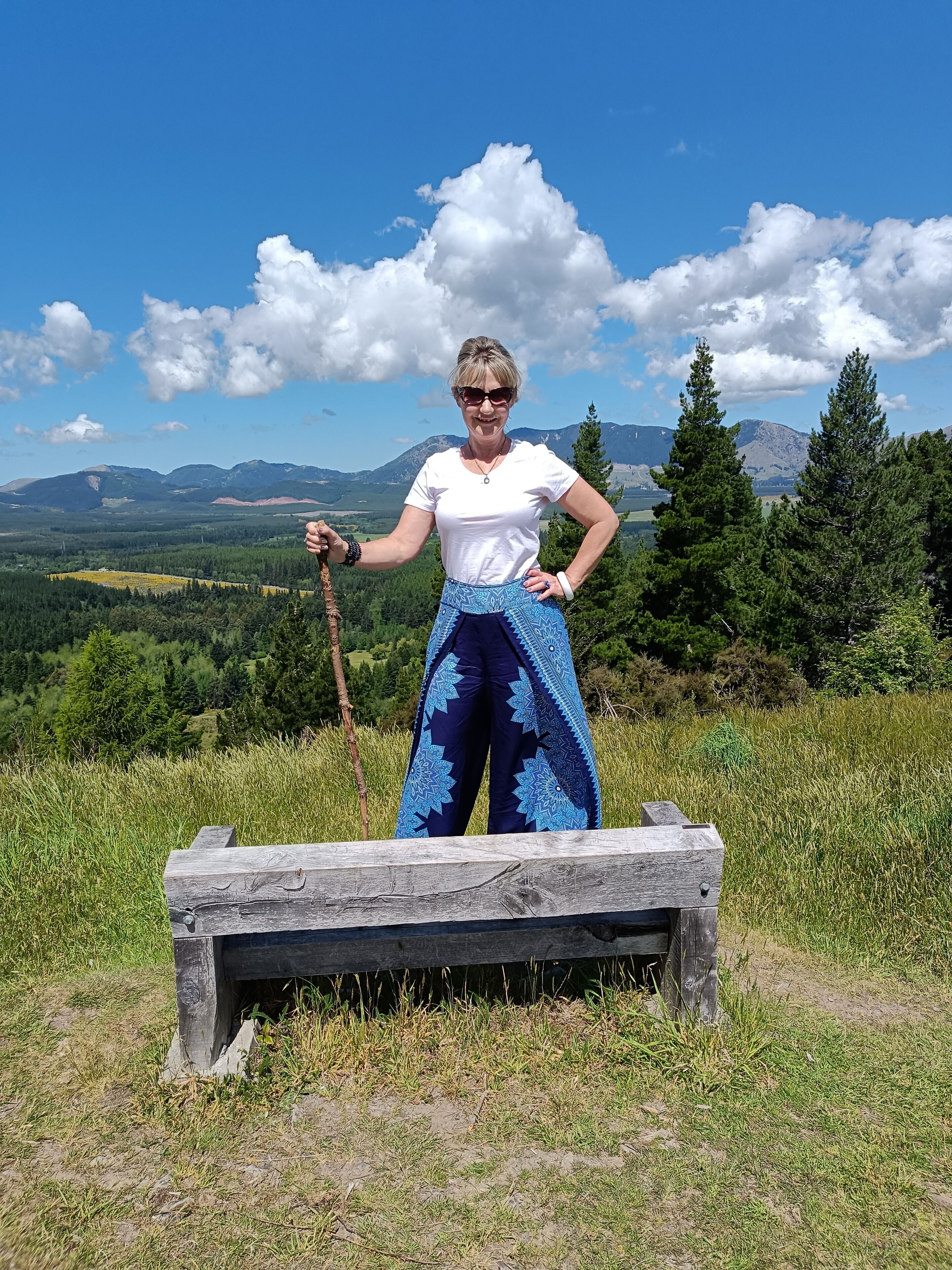 Una persona de pie en un banco de madera, con una vista panorámica de montañas y árboles al fondo.