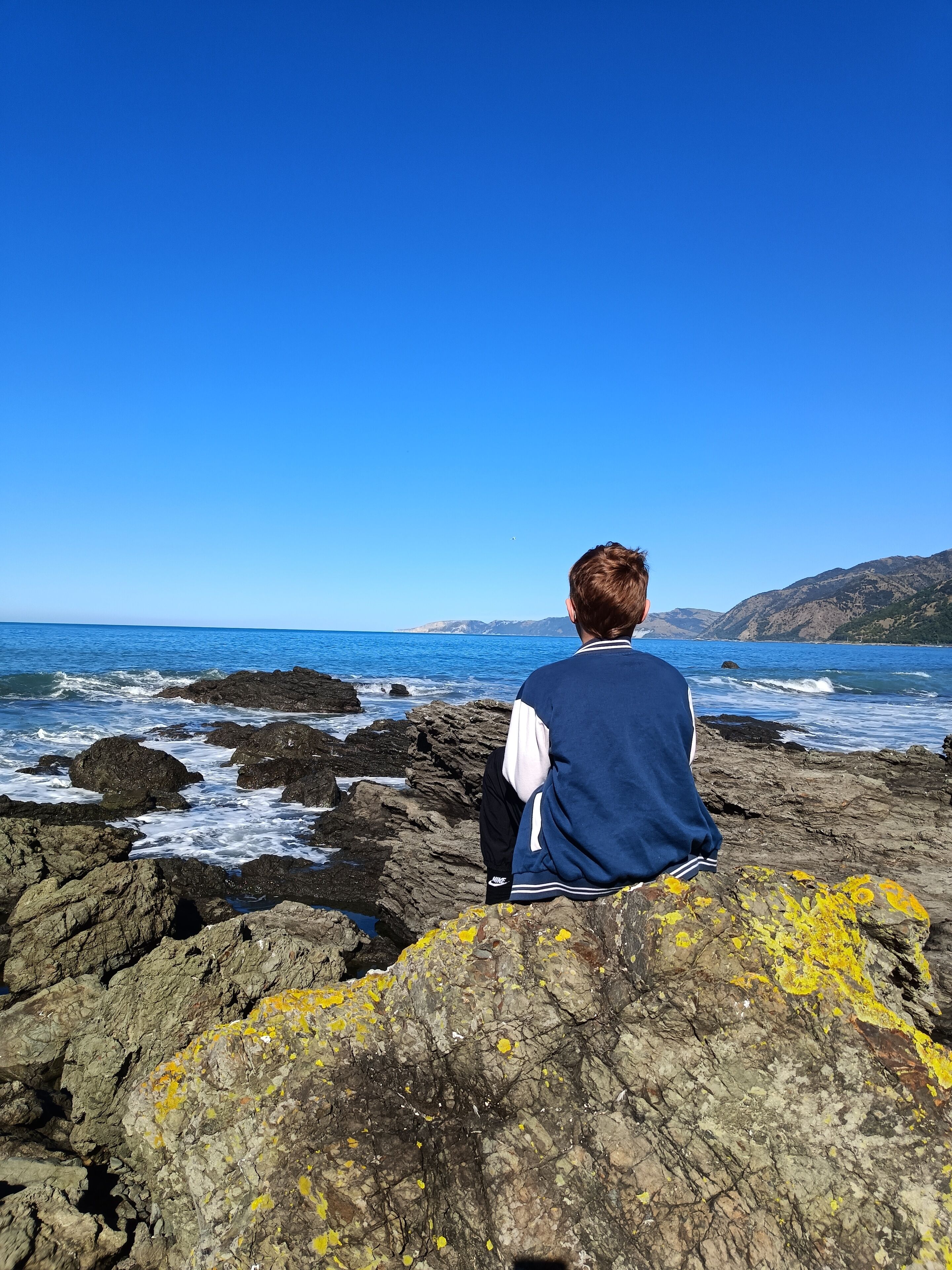 Una persona sentada sobre un saliente rocoso con vistas a la costa, donde las olas rompen contra las rocas.