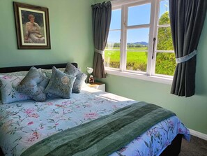 A bedroom with a bed, floral bedding, a nightstand, a framed picture, and a window with curtains. -  Country Cottage Delight in the Hurunui.
(Spotswood/Cheviot)
 (Spotswood)