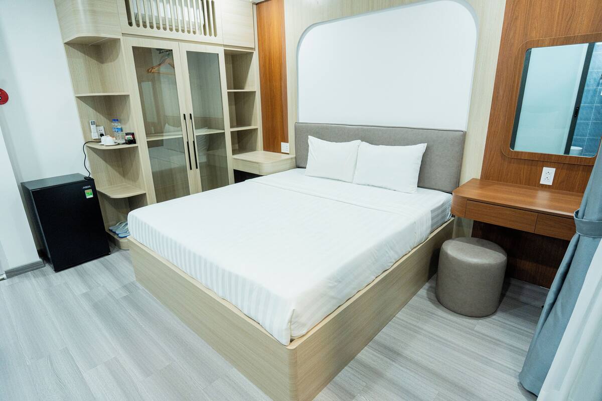 Une chambre d’hôtel bien rangée, avec un lit, une petite table de chevet, un miroir, une armoire et une poubelle.