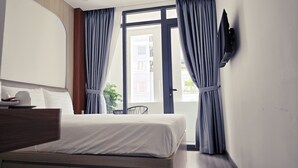 A hotel room with a bed, a desk, a chair, a TV mounted on the wall, and a window with curtains. - Mother's Home (Ho Chi Minh City)