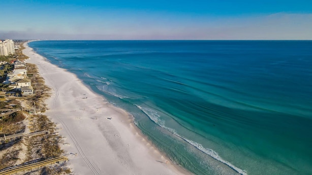 An aerial view of a beach with a long stretch of sandy shore, a few buildings, and a clear blue ocean.