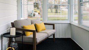 A cozy room with a wooden ceiling, a grey sofa with yellow cushions, a small black side table, and a view of the outdoors through large windows. - SquirrelHaus PEC Summ (Prince Edward)