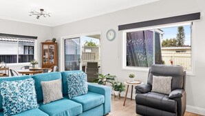 A modern living room with a teal sofa, a black leather chair, a ceiling fan, and a view of the outside through a large window. - Cottage by the Lake, 1 bedroom full size house, full kitchen, 2 living areas.  (Long Jetty)