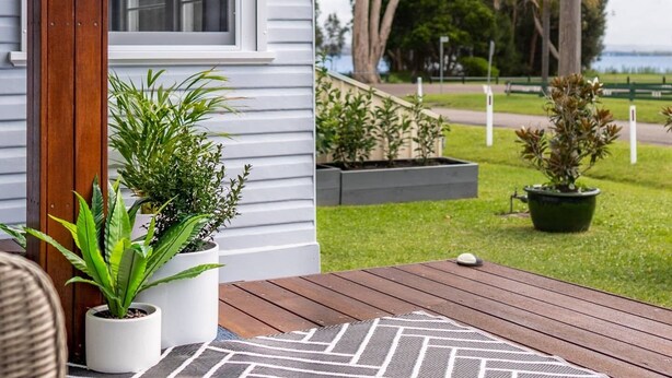 A wooden deck with potted plants, a window, and a view of a grassy area with trees and a fence. - Cottage by the Lake, 1 bedroom full size house, full kitchen, 2 living areas.  (Long Jetty)