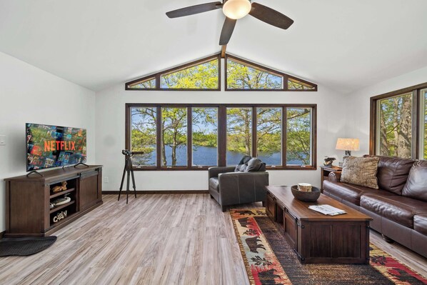 A spacious living room with a large window, a ceiling fan, a leather sofa, and a TV on a wooden stand.
