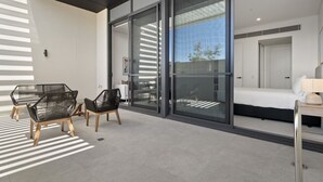 Una habitación de hotel moderna con un gran ventanal corredero que da a un balcón, una cama, una mesita de noche con lámpara y dos sillones de mimbre negros.