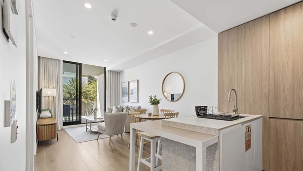 A modern kitchen with a central island, a dining area with a table and chairs, and a large sliding glass door leading to a balcony with a view of palm trees.