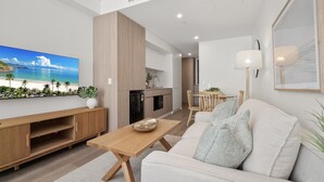Uma sala de estar moderna com sofá, mesa de centro e TV. Há uma área de cozinha com geladeira e mesa de jantar.