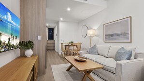 Uma sala de estar moderna com uma mesa de centro de madeira, um sofá cinza com almofadas, uma área de jantar com cadeiras de madeira e um quadro grande na parede.