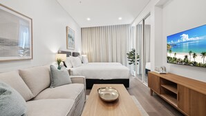 Quarto de hotel moderno com cama, sofá, mesa de centro, televisão e uma janela grande com cortinas.