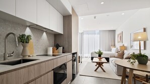 A modern kitchen with a built-in oven and a sink, a dining area with a table and chairs, and a living area with a sofa and a lamp. - Bright & Cosy Studio in Prime Fremantle Location (North Fremantle)