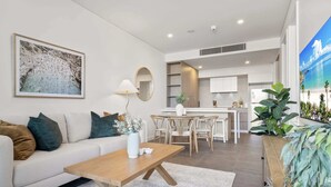 Un salon moderne avec un canapé, une table basse et un coin repas avec des chaises. On aperçoit au fond une cuisine équipée de placards et d’un évier.