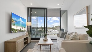 Una sala de estar moderna con un sofá, una mesa de centro y un televisor montado en la pared. Hay un balcón con vistas a la ciudad y una planta en el alféizar de la ventana.