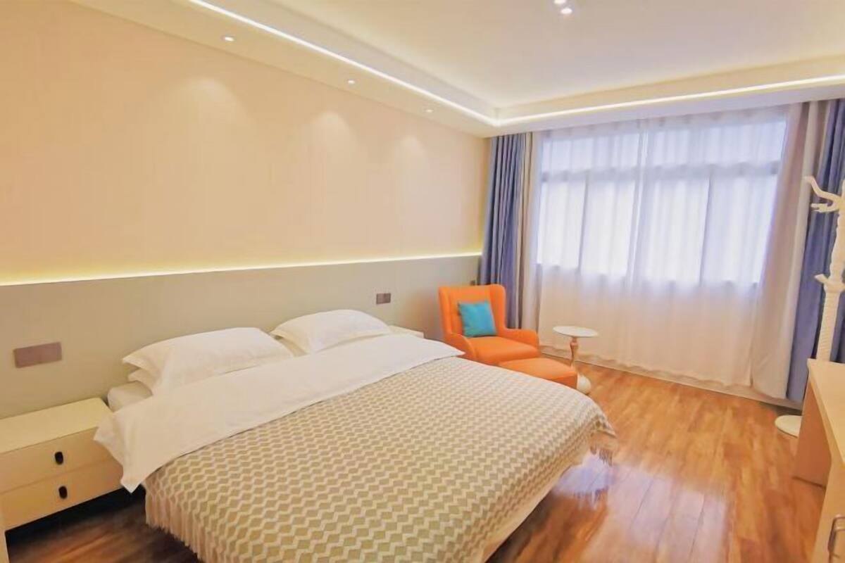 Quarto de hotel com uma cama grande, uma mesa de cabeceira, uma poltrona laranja, uma pequena mesa e uma janela ampla com cortinas.