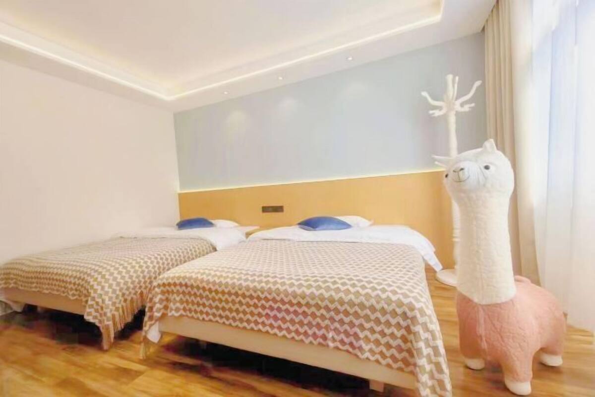 Quarto de hotel com duas camas, uma parede branca, luminária no teto e um bicho de pelúcia no chão.