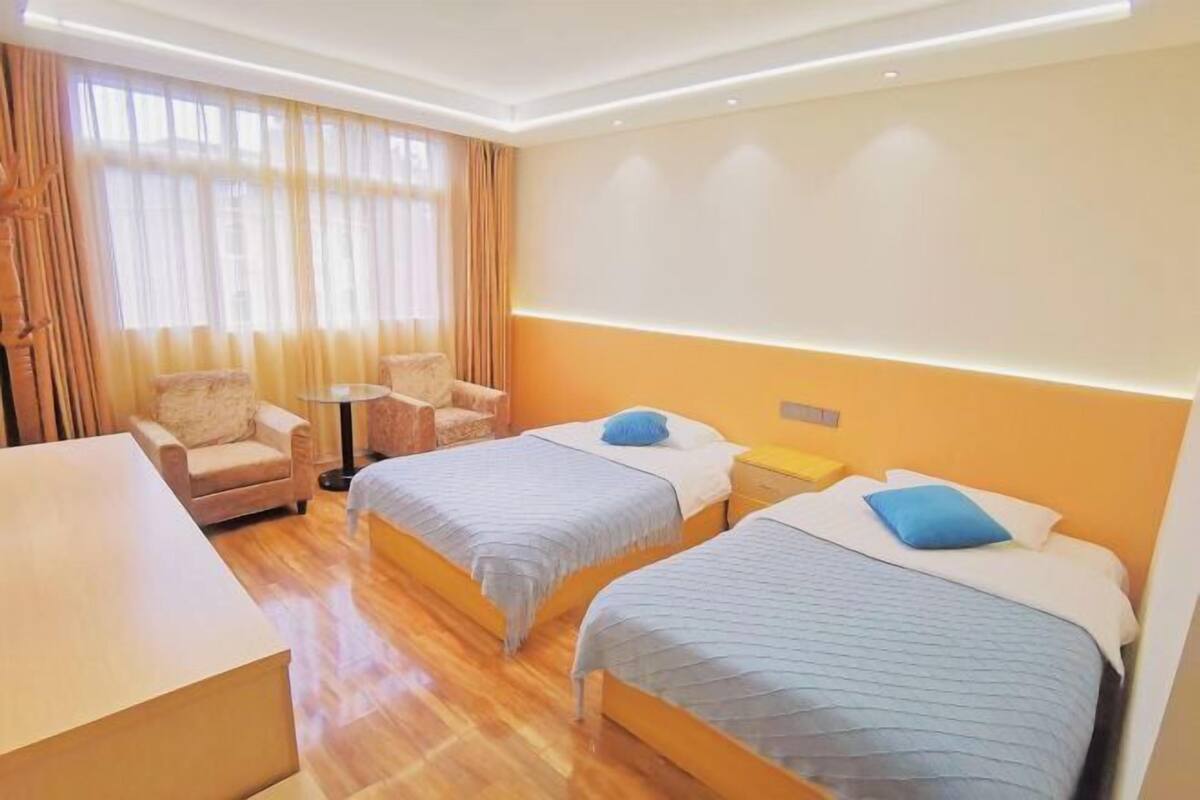 Quarto de hotel com duas camas, uma escrivaninha, uma cadeira e uma janela grande com cortinas.