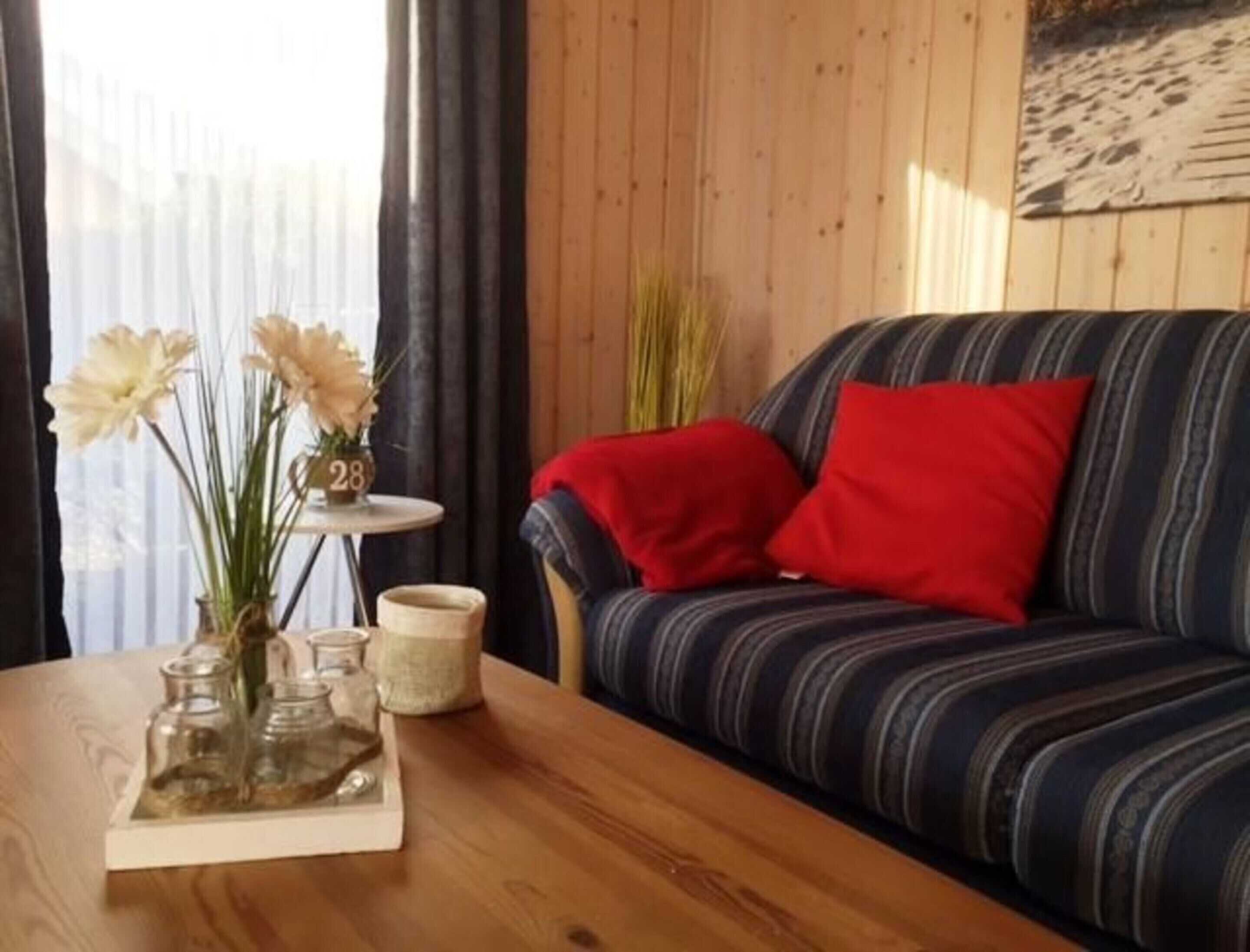 A wooden table with a vase of flowers, a small clock, and a candle. A striped sofa with red pillows is against a wooden wall.
