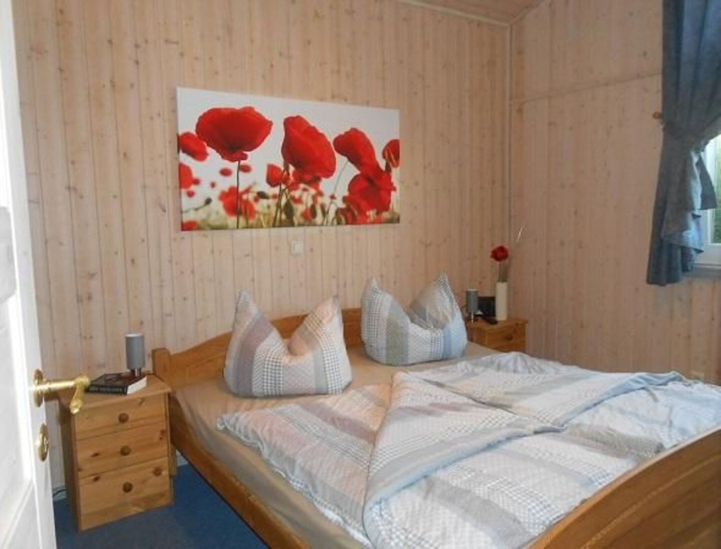 A bedroom with a wooden bed, a nightstand, a drawer unit, and a painting of red flowers on the wall.